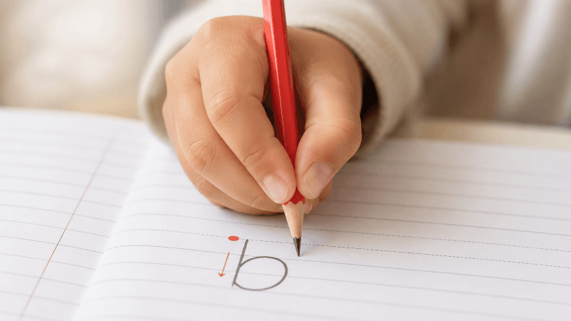 A child holding a red pencil in a correct tripod grip writing the lower-case letter b on ruled exercise-book paper